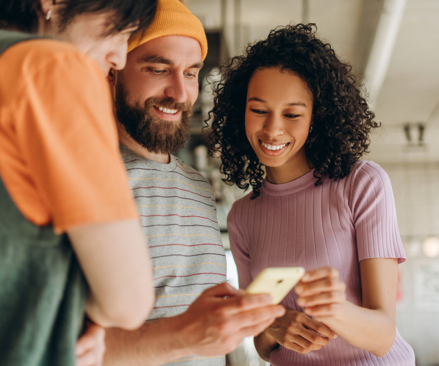 Three young colleagues smiling and collaborating on a project, sharing ideas while looking at a smartphone in a modern office filled with creativity and innovation. Online technology concept